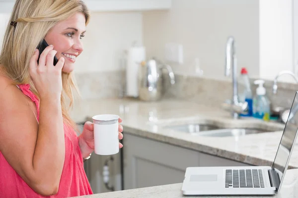 Gelukkige vrouw met laptop en koffie cup op oproep in de keuken — Stockfoto