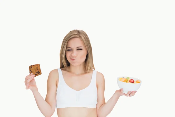 Woman with fruit bowl making faces as she looks at cookie — Stock Photo, Image