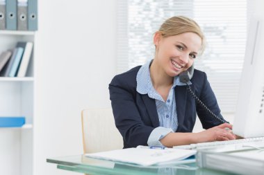 Portrait of business woman using phone and computer at desk clipart