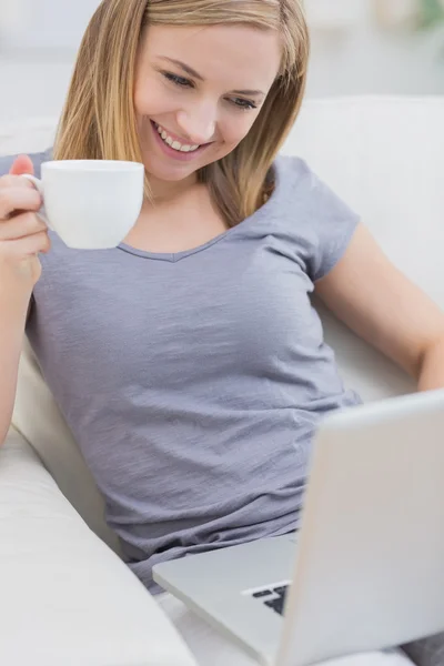 Mujer sonriente con taza de café usando computadora portátil en el sofá —  Fotos de Stock