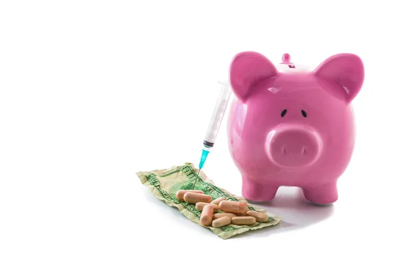 Syringe leaning against piggy bank with dollars and pills — Stock Photo, Image