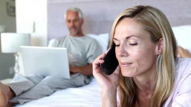 Vrouw lachen terwijl ze op de telefoon naast zijn echtgenoot op zijn laptop — Stockvideo