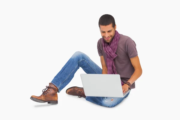 Χαμογελαστός άνθρωπος φορώντας κασκόλ που κάθεται στο πάτωμα χρησιμοποιώντας φορητό υπολογιστή — Φωτογραφία Αρχείου