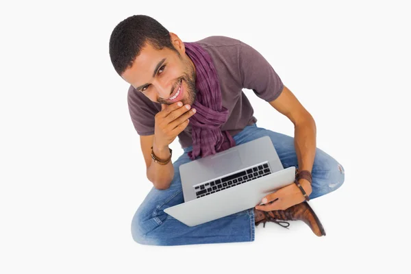 Hombre reflexivo sentado en el suelo usando el ordenador portátil y sonriendo a la cámara — Foto de Stock