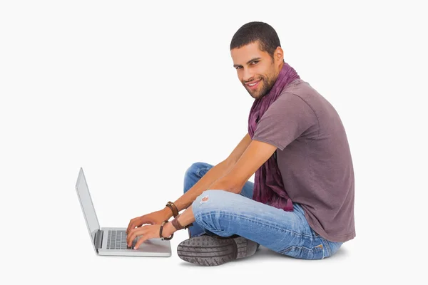 Модный мужчина, сидящий на полу с ноутбуком, улыбается перед камерой — стоковое фото