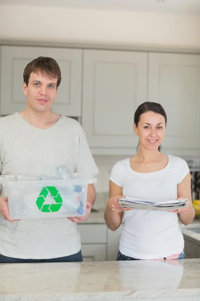 Smiling couple holding newspapers and recycling bin — Stock Photo, Image