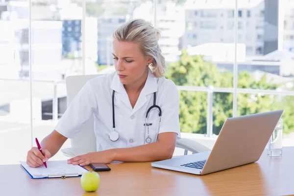 Blonde Krankenschwester schreibt auf einen Notizblock auf ihrem Schreibtisch — Stockfoto