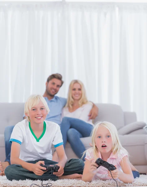 Los niños en la alfombra jugando videojuegos mientras sus padres un —  Fotos de Stock