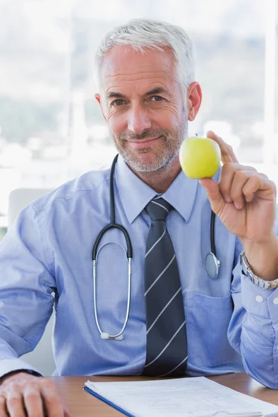 डॉक्टर अपने डेस्क के पीछे एक सेब पकड़े हुए बैठे — स्टॉक फ़ोटो, इमेज