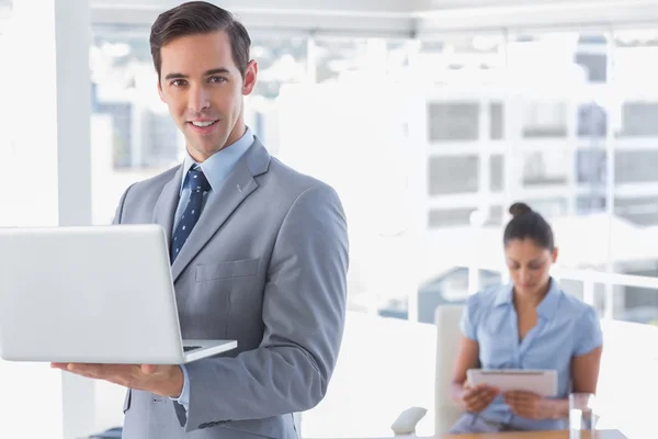 Empresário de pé com laptop e sorrindo para a câmera — Fotografia de Stock