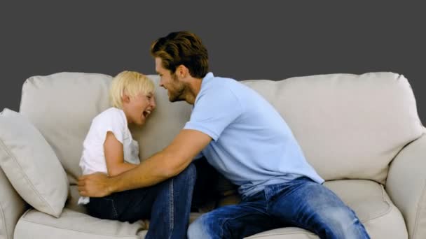 Padre haciendo cosquillas a su hijo en el sofá sobre fondo gris — Vídeo de stock