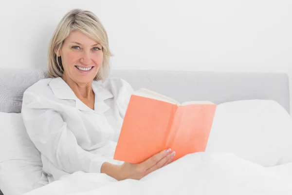 Blonde woman sitting in bed reading smiling at camera — Stock Photo, Image