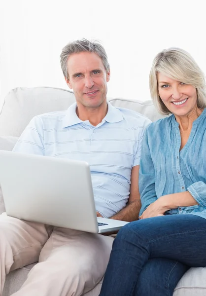 夫妇在相机微笑坐在沙发上一起使用的笔记本电脑 — 图库照片