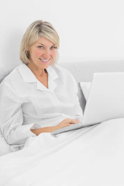 Femme blonde assise au lit avec un ordinateur portable souriant à la caméra — Photo