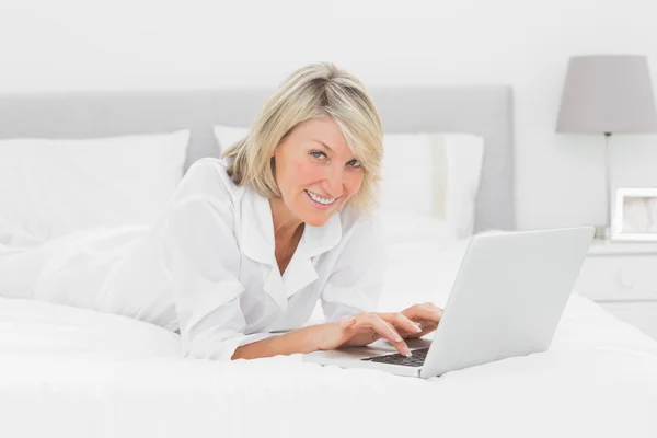 Sorrindo mulher usando seu laptop em sua cama olhando para a câmera — Fotografia de Stock