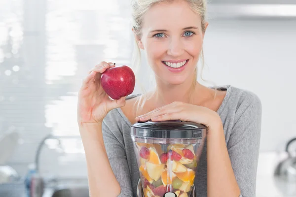 Blonde vrouw leunend op haar juicer vol met fruit en rood te houden — Stockfoto