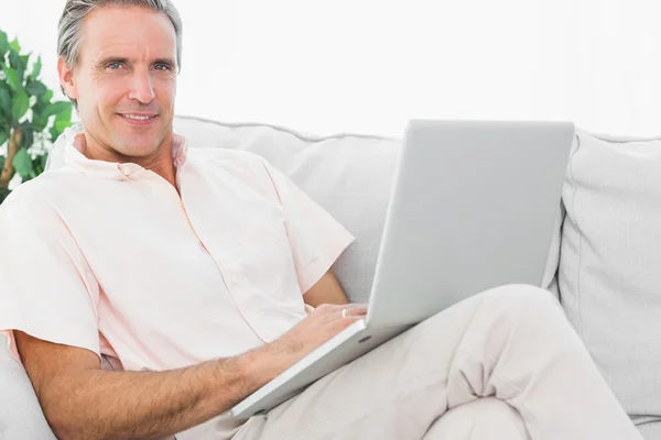 Homem alegre em seu sofá usando laptop olhando para a câmera — Fotografia de Stock