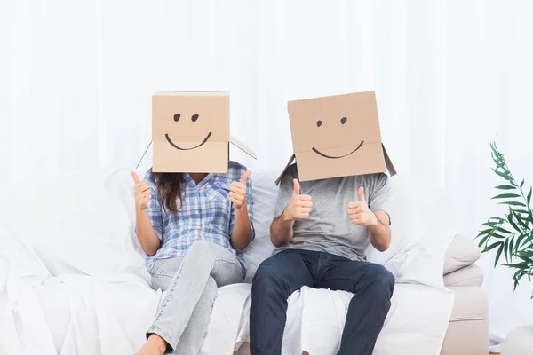 Couple sitting with cardboard boxes on head giving thumbs up — Stock Photo, Image