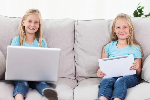 Twins using a laptop and a tablet sitting on a couch — Stock Photo, Image