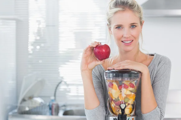 Gelukkige vrouw leunend op haar juicer vol met fruit en rood te houden — Stockfoto