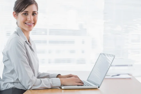 Mujer de negocios escribiendo en su portátil en el escritorio y sonriendo a la cámara —  Fotos de Stock