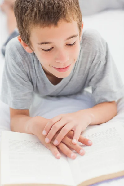 Smiling little boy lying on bed reading book — Stock Photo, Image