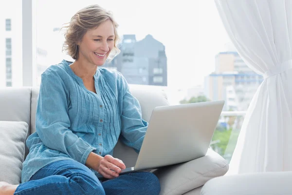 快活的金发女人坐在她的沙发使用的便携式计算机上 — 图库照片