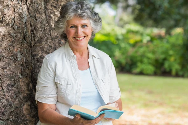 Cheerful mature woman holding book sitting on tree trunk — Stock Photo, Image