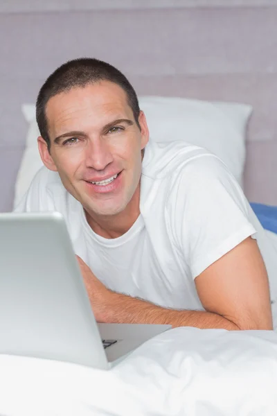 Cheerful man lying on bed using his laptop — Stock Photo, Image