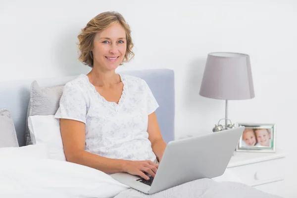 Zufriedene blonde Frau sitzt mit Laptop im Bett — Stockfoto