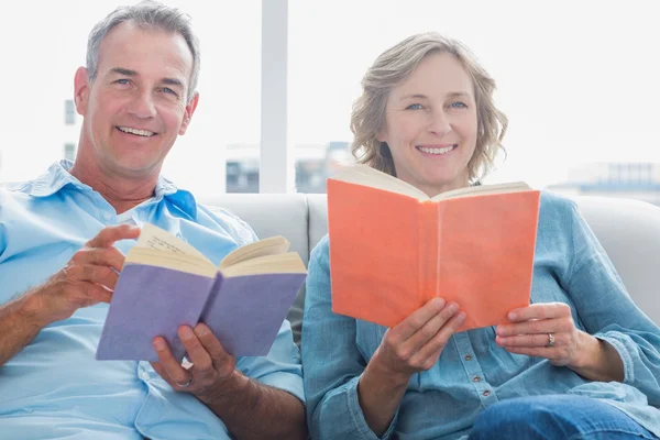 Relajada pareja leyendo libros en el sofá sonriendo a la cámara —  Fotos de Stock