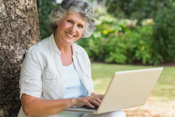 Ağaç gövdesi üzerinde oturan bir dizüstü bilgisayar kullanarak neşeli olgun kadın — Stok fotoğraf