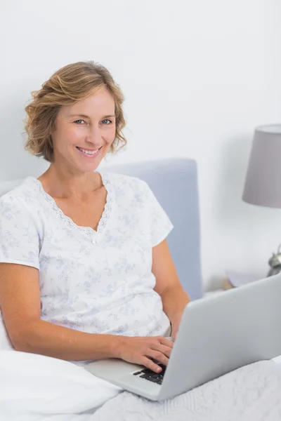 Smiling blonde woman sitting in bed using laptop — Stock Photo, Image