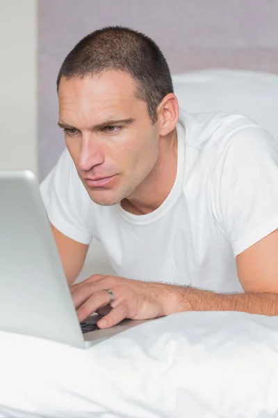 Серйозний чоловік лежить на ліжку, використовуючи свій ноутбук — стокове фото
