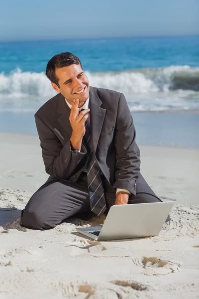 Улыбающийся бизнесмен сидит на песке со своим ноутбуком — стоковое фото