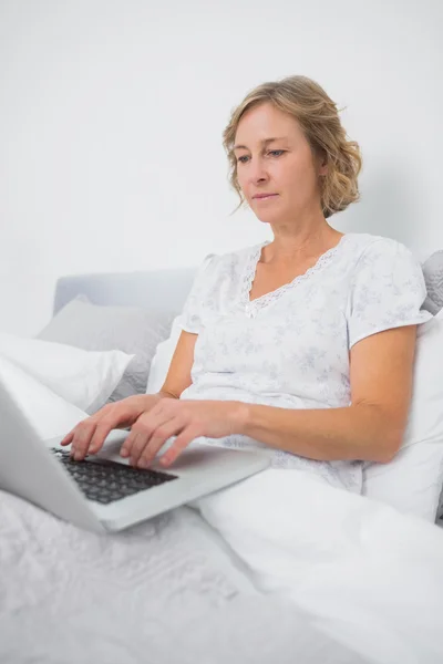 Relajada mujer rubia sentada en la cama usando el ordenador portátil — Foto de Stock