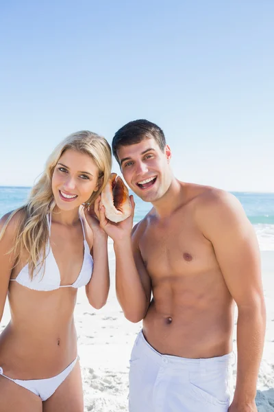 Attractive couple listening to seashell smiling at camera — Stock Photo, Image