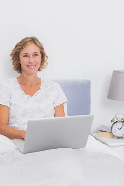 微笑着的金发女人坐在床上使用笔记本电脑 — 图库照片