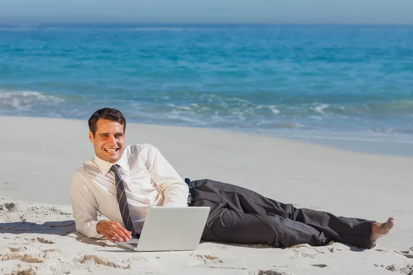 Jovem empresário sorridente deitado na areia com seu laptop — Fotografia de Stock