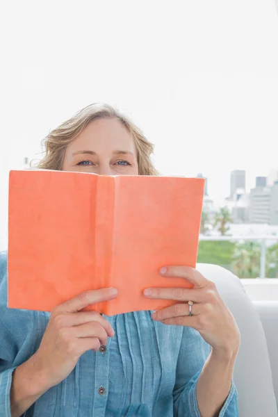 Frau sitzt auf ihrem Sofa und bedeckt Gesicht mit orangefarbenem Buch — Stockfoto