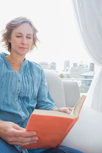 Innehåll blond kvinna sitter på hennes soffa läser en bok — Stockfoto