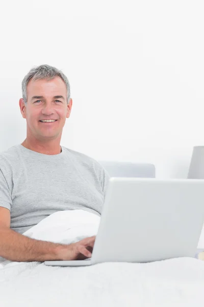 Allegro uomo dai capelli grigi che usa il suo computer portatile a letto — Foto Stock