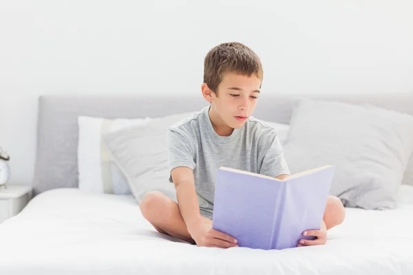 Koncentrált kisfiú ül könyvével ágyon — Stock Fotó