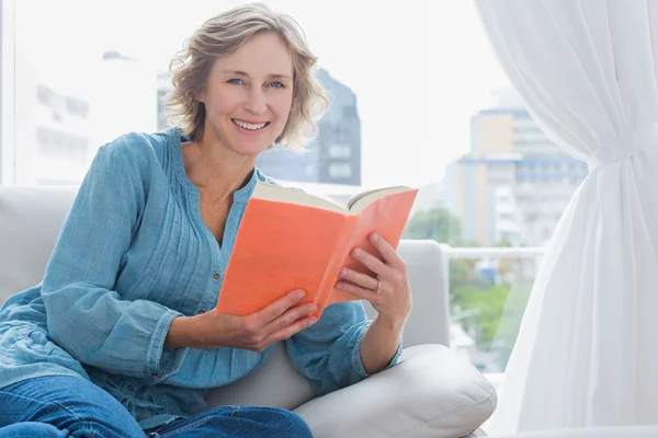 Fröhliche blonde Frau sitzt auf ihrem Sofa und hält ein Buch — Stockfoto