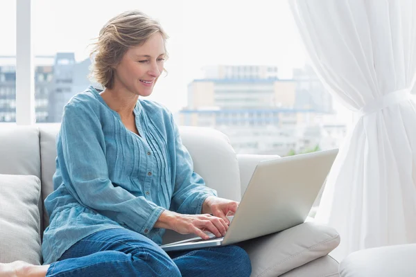 Glada blonda kvinnan sitter på hennes soffa med laptop — Stockfoto