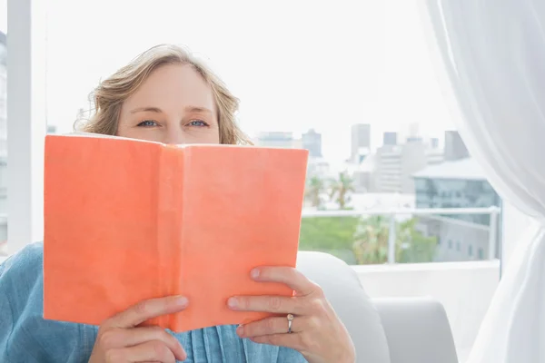 Blonde woman sitting on her couch covering face with orange book — Stock Photo, Image