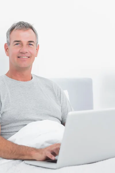 Sorrindo homem de cabelos grisalhos usando seu laptop na cama — Fotografia de Stock