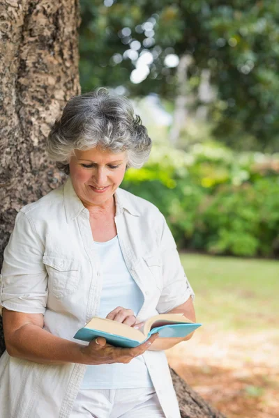 Cheerful mature woman reading book leaning on tree trunk — Stock Photo, Image