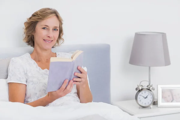 Mujer rubia feliz sentada en la cama sosteniendo libro —  Fotos de Stock