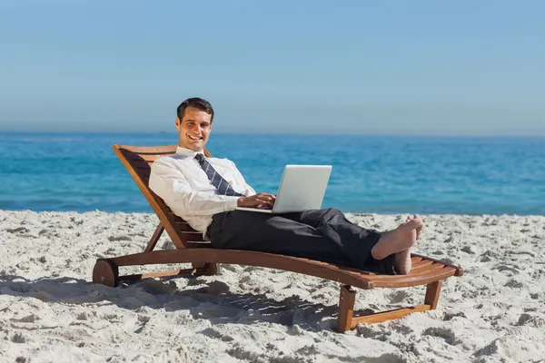 Jovem empresário sorridente deitado em uma cadeira de praia com seu laptop — Fotografia de Stock